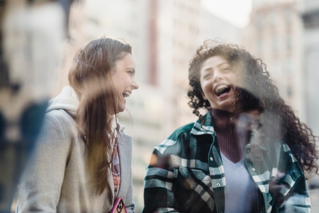 Two women, seen through a window, stand together on a city street and laugh