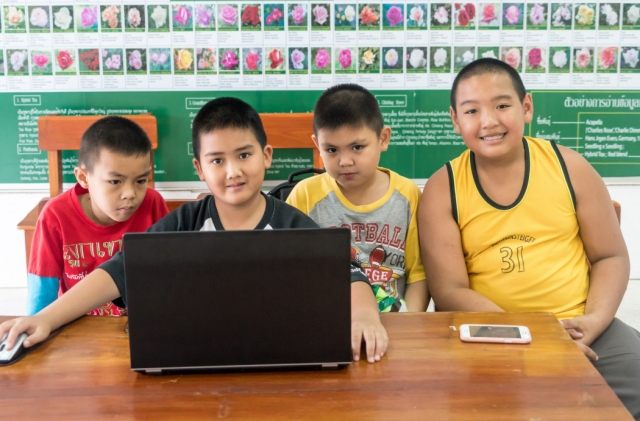 Four young boys in a classroom sit at a table with a laptop and smart phone