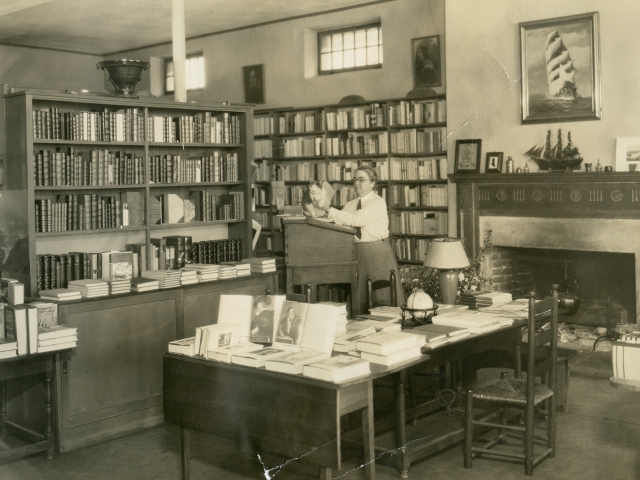 Aged photograph showing Marion E. Dodd and her cat surrounded by bookshelves and display tables in a large room at The Hampshire Bookshop