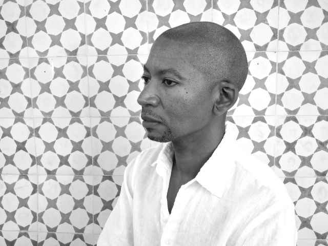 Photographic portrait of the poet Myronn Hardy. Hardy is shown in near-profile in a white shirt with patterned tiles in the background.