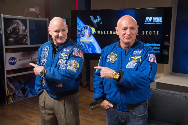 Photograph of twins Scott and Mark Kelly in the NASA twin study. The twins wear matching NASA jackets and both point to their left.