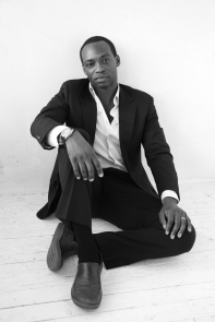 A Black man seated, one leg crossed over the other, wearing a suit.